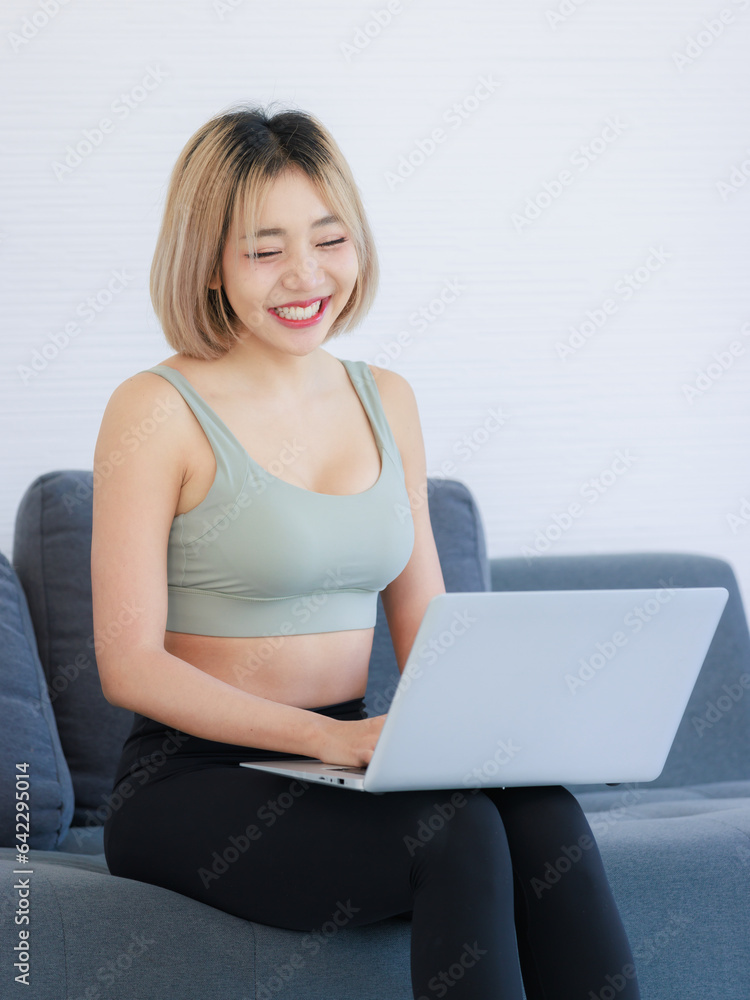 Asian young fit sexy female teenager in sportswear sport bra legging sneakers sitting on cozy sofa couch on call with smartphone surfing browsing internet online via laptop notebook computer at home Asian young fit sexy female teenager in sportswear sport bra legging sneakers sitting on cozy sofa couch on call with smartphone surfing browsing internet online via laptop notebook computer at home