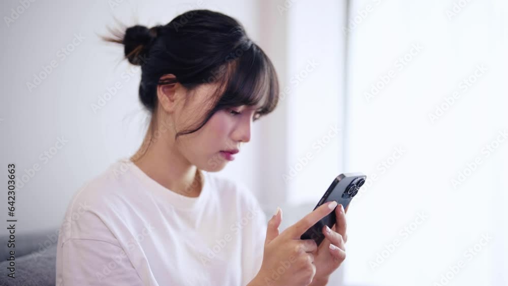 Millennial asian young woman relax on comfortable couch at home chatting with her friends and scroll through social media on her phone. Millennial asian young woman relax on comfortable couch at home chatting with her friends and scroll through social media on her phone.