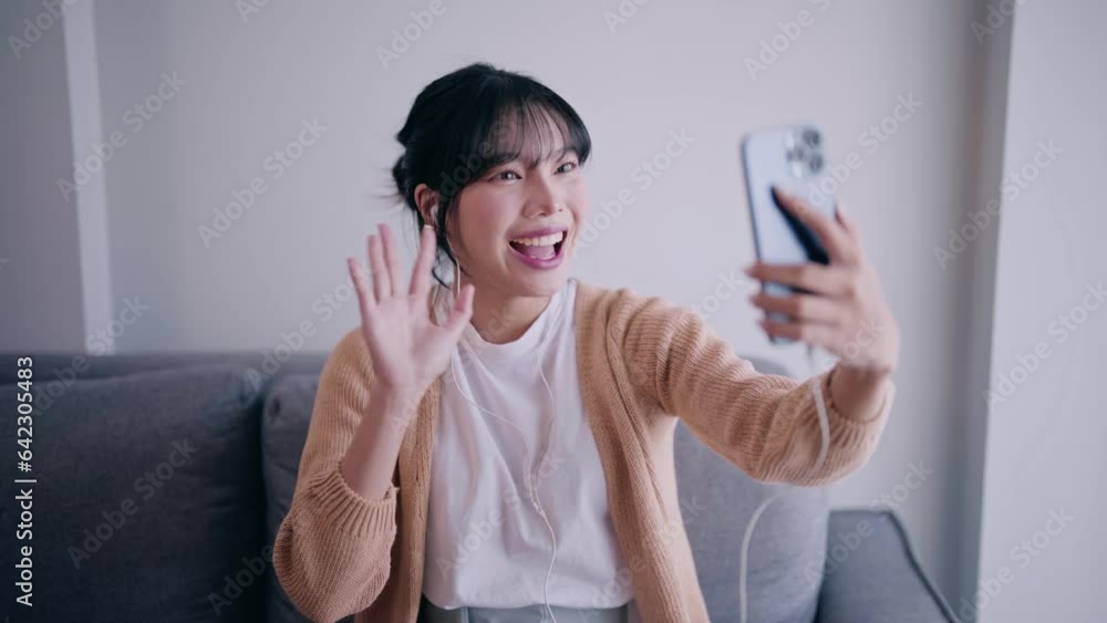 Happy young asian woman having video call and waving hand to friend on mobile cell phone or smartphone in living room relaxing at home. Happy young asian woman having video call and waving hand to friend on mobile cell phone or smartphone in living room relaxing at home.