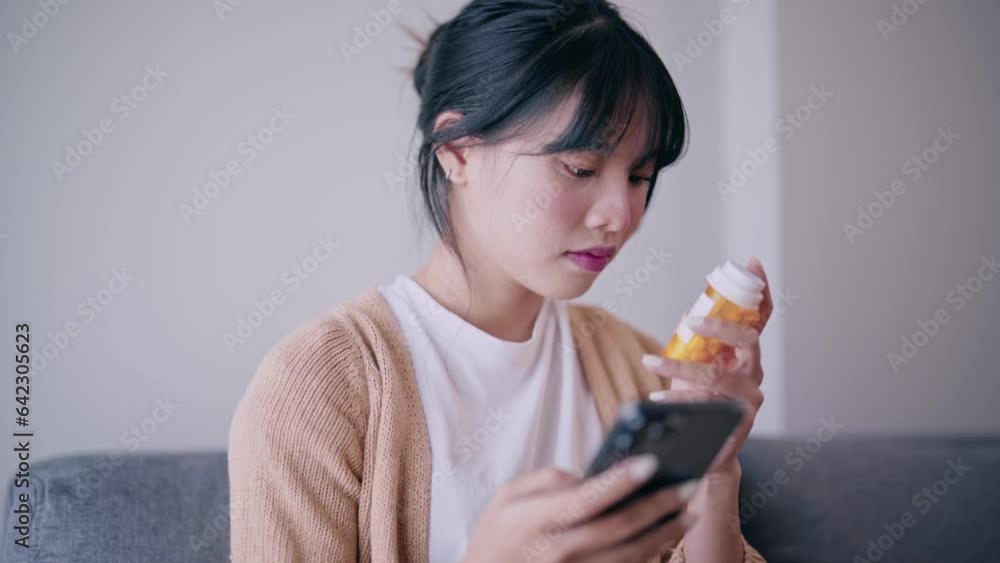 Worried Asian girl check a bottle of drug and searching information about medicines on smartphone take medicine after doctor order sitting on a couch in the living room in a house. Worried Asian girl check a bottle of drug and searching information about medicines on smartphone take medicine after doctor order sitting on a couch in the living room in a house.