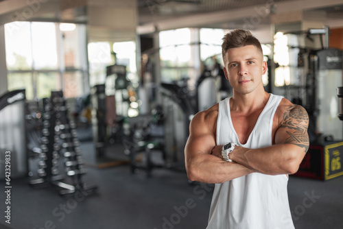 Cool handsome muscular healthy athletic man with a hairstyle with a tattoo on his arm in a white tank top stands hand in hand in the sport gym
