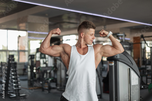 Handsome strong muscular man athlete with hairstyle in white tank top with biceps shows muscles in the sport gym