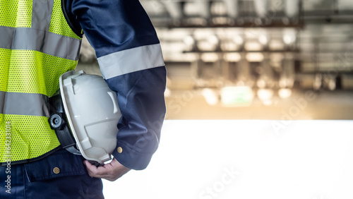 Safety workwear concept. Male hand holding white safety helmet or hard hat. Construction worker man in protective suit and reflective green vest standing with building piping system in the background