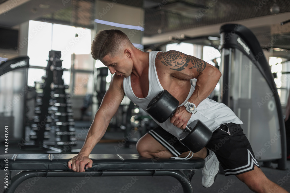 Strong handsome athletic man with a muscular body stands near the bench and does an exercise with dumbbells in the gym Strong handsome athletic man with a muscular body stands near the bench and does an exercise with dumbbells in the gym