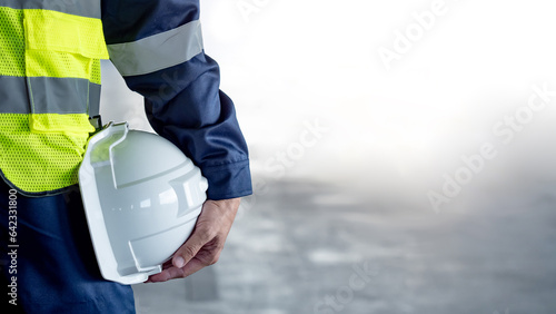 Safety workwear concept. Male hand holding white safety helmet or hard hat. Construction worker man in protective suit and reflective green vest standing with building concrete floor in the background photo