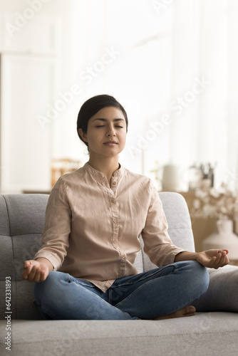 Peaceful young Indian yogi woman training focus, concentration, meditating sofa on short break, sitting in lotus yoga pose, keeping zen mudra, taking deep breath with closed eyes