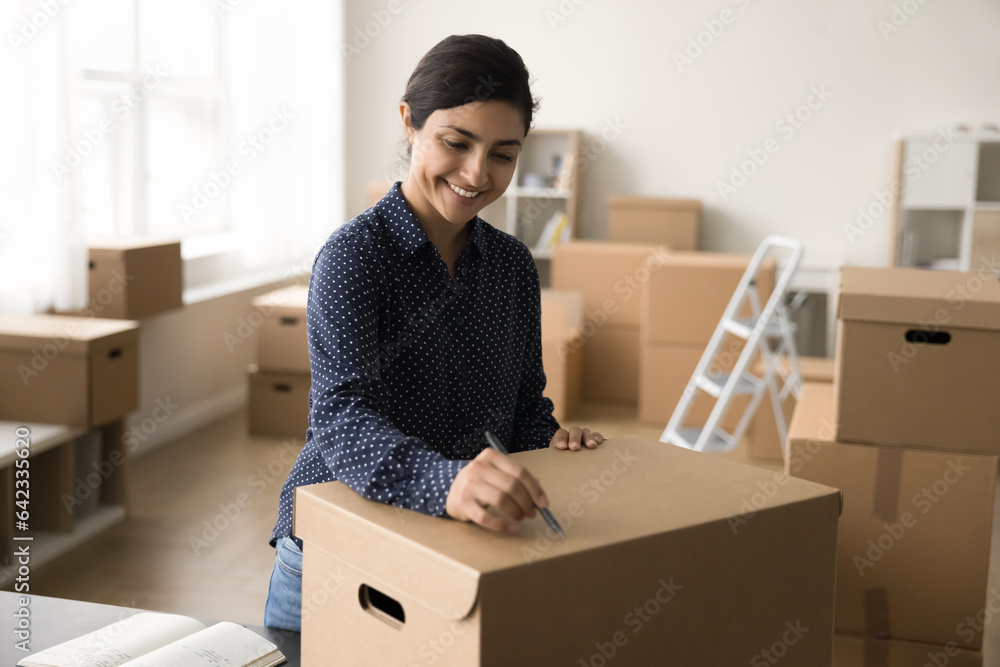 Positive beautiful Indian online shop manager preparing purchase for sending, writing notes, address on closed packed cardboard box for shipping, smiling, laughing, enjoying business process Positive beautiful Indian online shop manager preparing purchase for sending, writing notes, address on closed packed cardboard box for shipping, smiling, laughing, enjoying business process