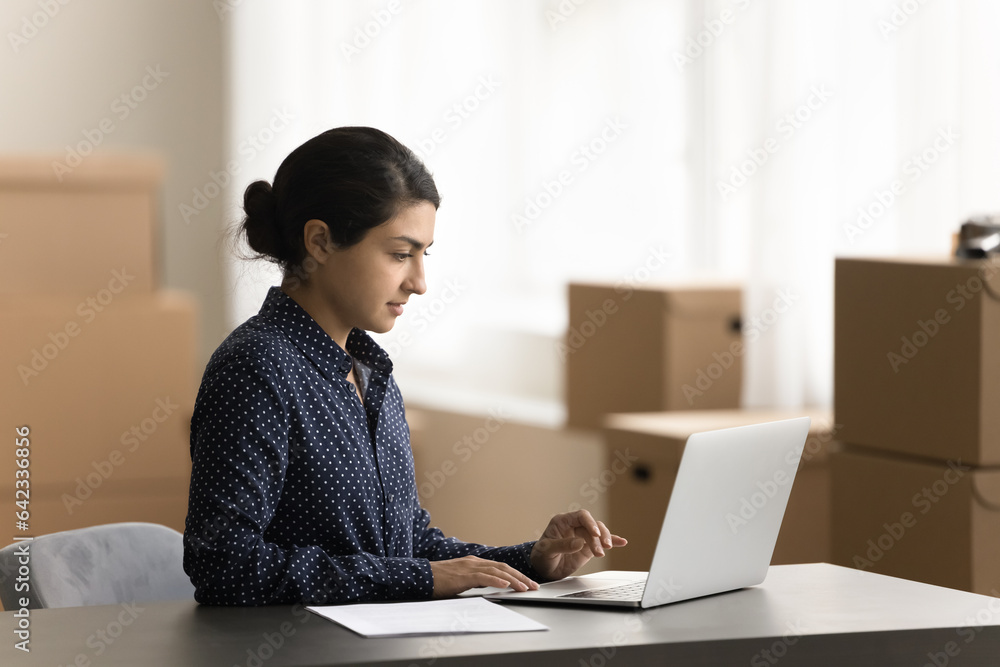 Focused pretty Indian Internet store employee woman working at laptop in storage room with cardboard packages, transportation boxes in background, typing, processing order Focused pretty Indian Internet store employee woman working at laptop in storage room with cardboard packages, transportation boxes in background, typing, processing order