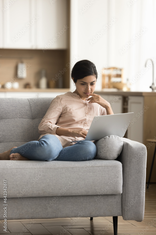Focused Indian freelance employee woman using professional online application on laptop computer on sofa at home, typing, touching chin, thinking on ideas for project, reading text Focused Indian freelance employee woman using professional online application on laptop computer on sofa at home, typing, touching chin, thinking on ideas for project, reading text