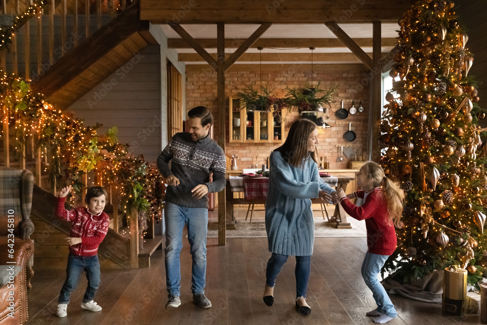 Happy young couple parents dancing to energetic disco music with adorable little children son daughter near Christmas tree in beautifully decorated house, New Year holidays celebration concept.