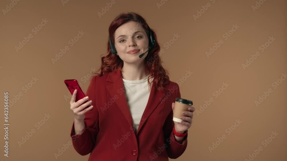 Medium isolated video of a happy and relaxed young woman in a call center headset holding a smartphone in one hand and a cup of coffee in the other. Medium isolated video of a happy and relaxed young woman in a call center headset holding a smartphone in one hand and a cup of coffee in the other.