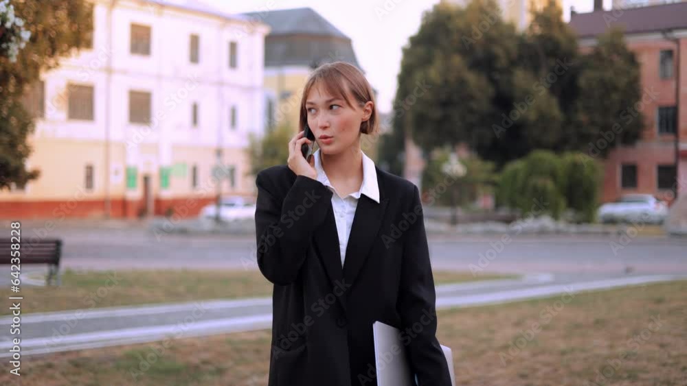 Portrait of a Caucasian young serious woman with short hair business lady standing outdoors on the street holding a closed laptop in her hand and talking on the mobile phone, looking sideways confiden Portrait of a Caucasian young serious woman with short hair business lady standing outdoors on the street holding a closed laptop in her hand and talking on the mobile phone, looking sideways confiden
