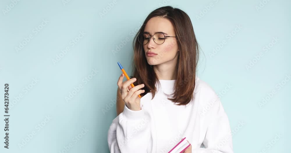 Young beautiful girl writing in pink diary her romantic story, poem while she is smiling, dreaming, thinking. Creative woman in eyeglasses writing with pen at her pink notebook on blue background. Young beautiful girl writing in pink diary her romantic story, poem while she is smiling, dreaming, thinking. Creative woman in eyeglasses writing with pen at her pink notebook on blue background.