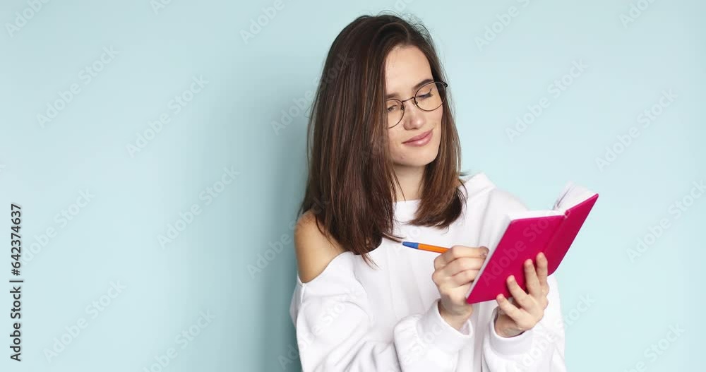 Young beautiful girl writing in pink diary her romantic story, poem while she is smiling, dreaming, thinking. Creative woman in eyeglasses writing with pen at her pink notebook on blue background. Young beautiful girl writing in pink diary her romantic story, poem while she is smiling, dreaming, thinking. Creative woman in eyeglasses writing with pen at her pink notebook on blue background.