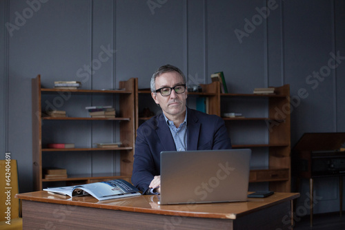 Portrait of mature serious businessman in suit and glasses pensivly doing his work on the laptop in office photo