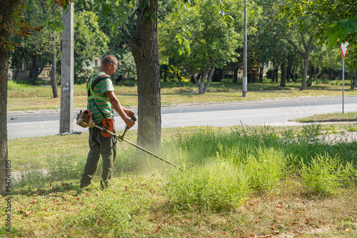 Man gardener mowing ragweed bushes, ambrosia artemisiifolia that causing allergy summer and autumnwith electric or petrol lawn trimmer along the city street. Gardening care tools and equipment.