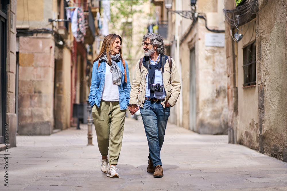 Happy adult Caucasian couple strolling hand in hand along beautiful tourist street in city. Mature people in love enjoying a romantic vacation walk. Positive relationships and weekend getaways spring.