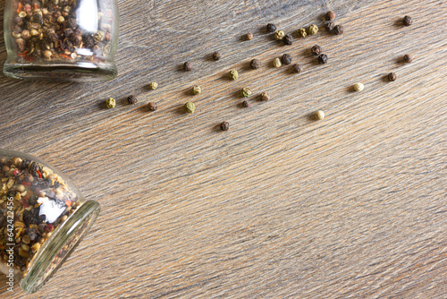 Peas of multi-colored peppers and glass pepper shakers are scattered on a wooden table.