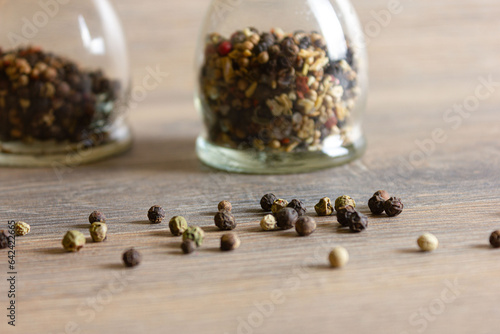 Peas of multi-colored peppers and glass pepper shakers are scattered on a wooden table.