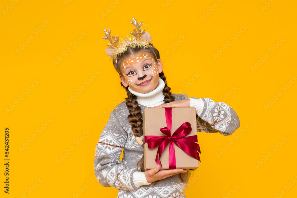 A little girl is dressed in a carnival festive deer with horns. Child holds a gift box with a red bow. New Year's holidays for children. Merry Christmas. Yellow isolated background.