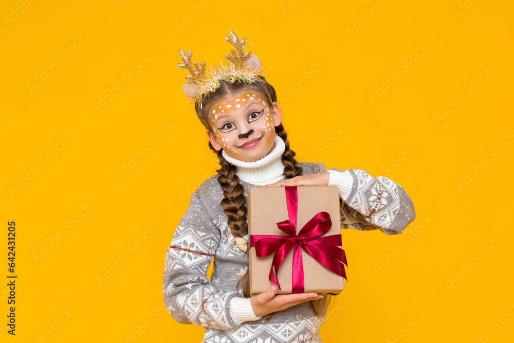 A little girl is dressed in a carnival festive deer with horns. Child holds a gift box with a red bow. New Year's holidays for children. Merry Christmas. Yellow isolated background. A little girl is dressed in a carnival festive deer with horns. Child holds a gift box with a red bow. New Year's holidays for children. Merry Christmas. Yellow isolated background.