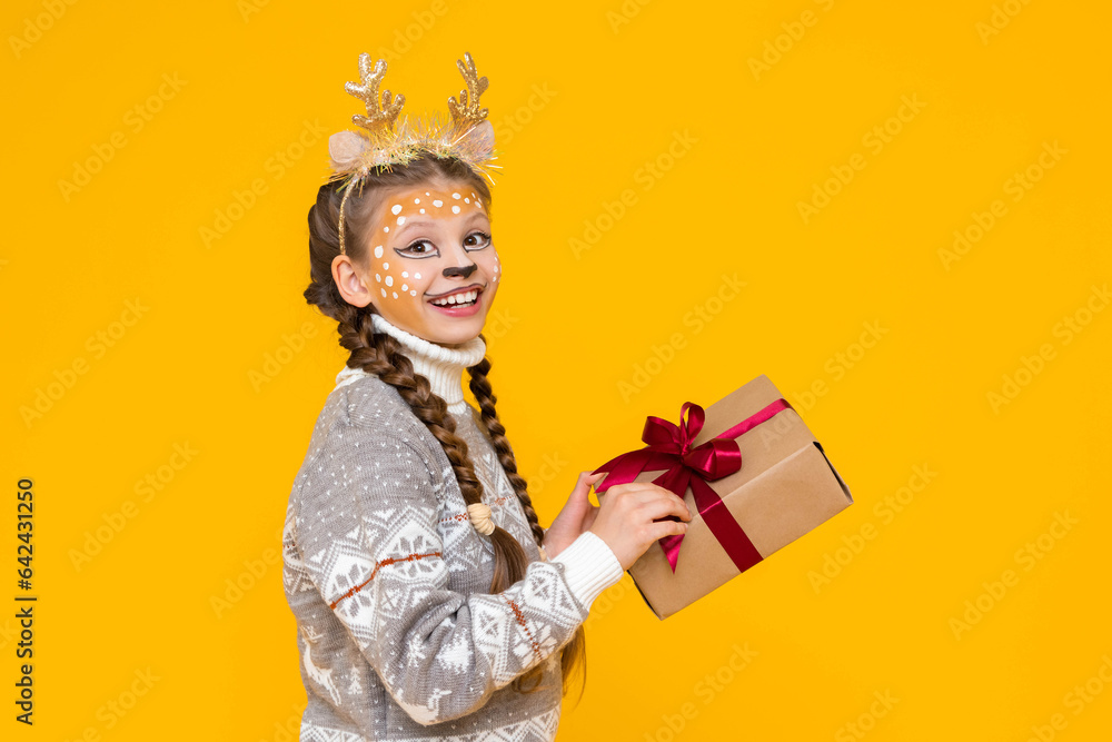 A little girl is dressed in a carnival festive deer with horns. Child holds a gift box with a red bow. New Year's holidays for children. Merry Christmas. Yellow isolated background.