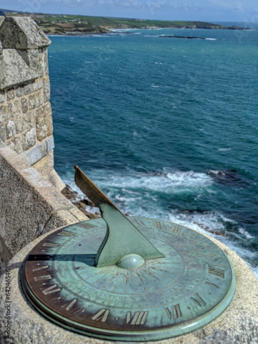 A metal sundial with roman numerals on a castle wall with the ocean behind it.