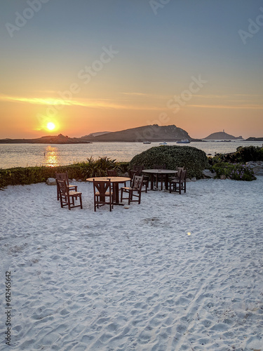 Two wooden tables with chairs on a white sandy beach by the ocean at sunset and islands and coastline visible on the horizon, taken on the island of St Martins, Isles of Scilly