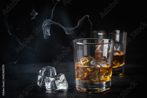 Whiskey with ice in glass and smoke on dark background, copy space