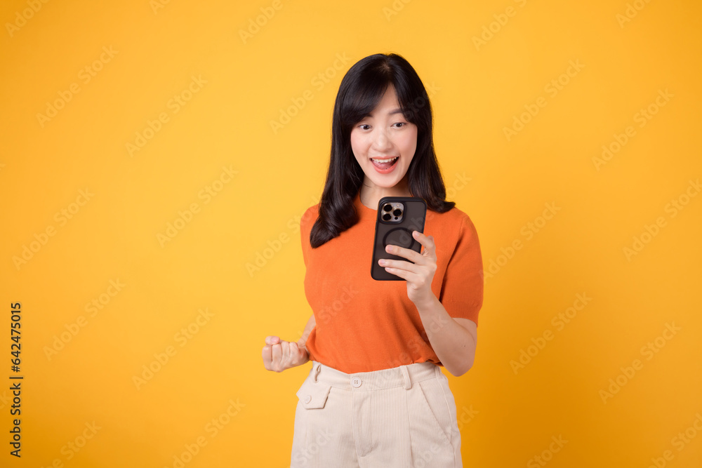 Smiling Asian woman in 30s, wearing orange shirt, using smartphone with fist up hand sign on vibrant yellow background. Celebrate new app concept