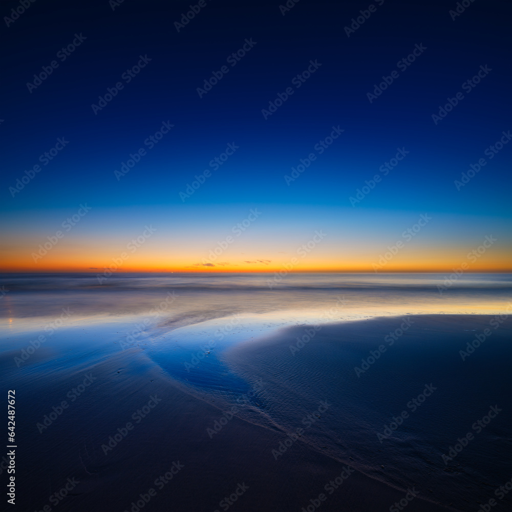 Fototapeta premium A seascape during sunset. Lines of sand on the seashore. Bright sky during sunset. A sandy beach at low tide. Wallpaper and background.