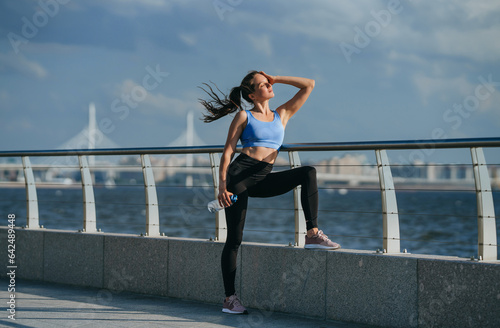 Tired American girl relaxing after hard workout outside standing at embankment eyes closed enjoying sun rays at sunset time against sea shore and bridge on background. Jogging, healthy people. photo