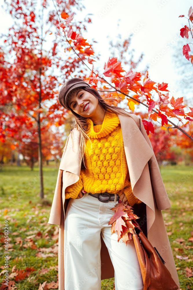Stylish woman wears yellow sweater holding handbag in autumn park enjoing landscape. Fall female clothes and accessories