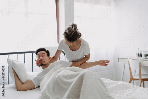 Funny asian couple wife try to wake lazy husband up in white bedroom.