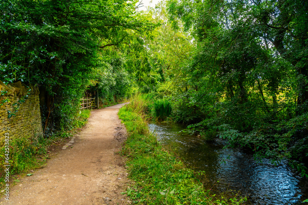 A scenic shady dirt path walkway along the River Eye in the English countryside at the Cotswold village of Lower Slaughter, Gloucestershire, England UK. A scenic shady dirt path walkway along the River Eye in the English countryside at the Cotswold village of Lower Slaughter, Gloucestershire, England UK.