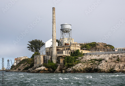 San Francisco, CA, USA - July 12, 2023: Alcatraz Island NE tip closeup with tall chimney, water tower.. White guano covered rocky cliffs, sparse green foliage, under light blue sky and greenish water