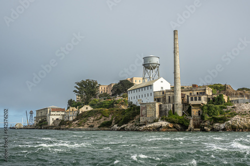 San Francisco, CA, USA - July 12, 2023: Alcatraz Island NE shoreline with tall chimney, water tower. Main building on top and east side under light gray sky with greenish bay water