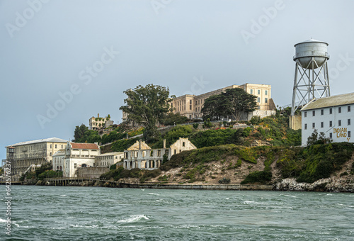 San Francisco, CA, USA - July 12, 2023: Alcatraz Island east shoreline with main prison building on top and other east side buildings plus water tower under light gray sky and greenish water.