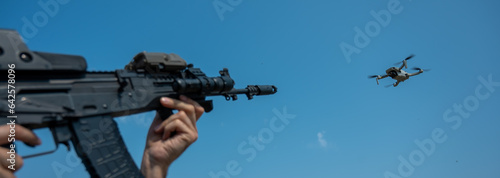 A man aims to shoot a rifle at a flying drone against a blue sky. 