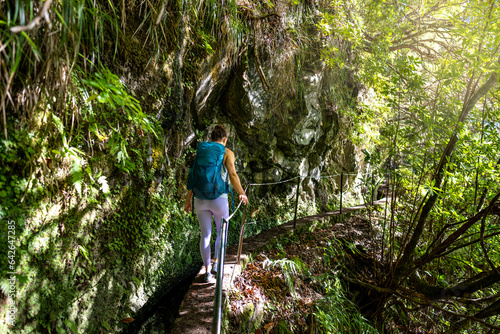 Backpacker toursit walking along sunny rainforst water channel trail under overgrown rock wall. Levada of Caldeirão Verde, Madeira Island, Portugal, Europe.