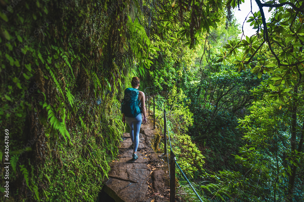 Female backpacker walking along rainforest water channel path on steep cliff covered with plants. Levada of Caldeirão Verde, Madeira Island, Portugal, Europe.