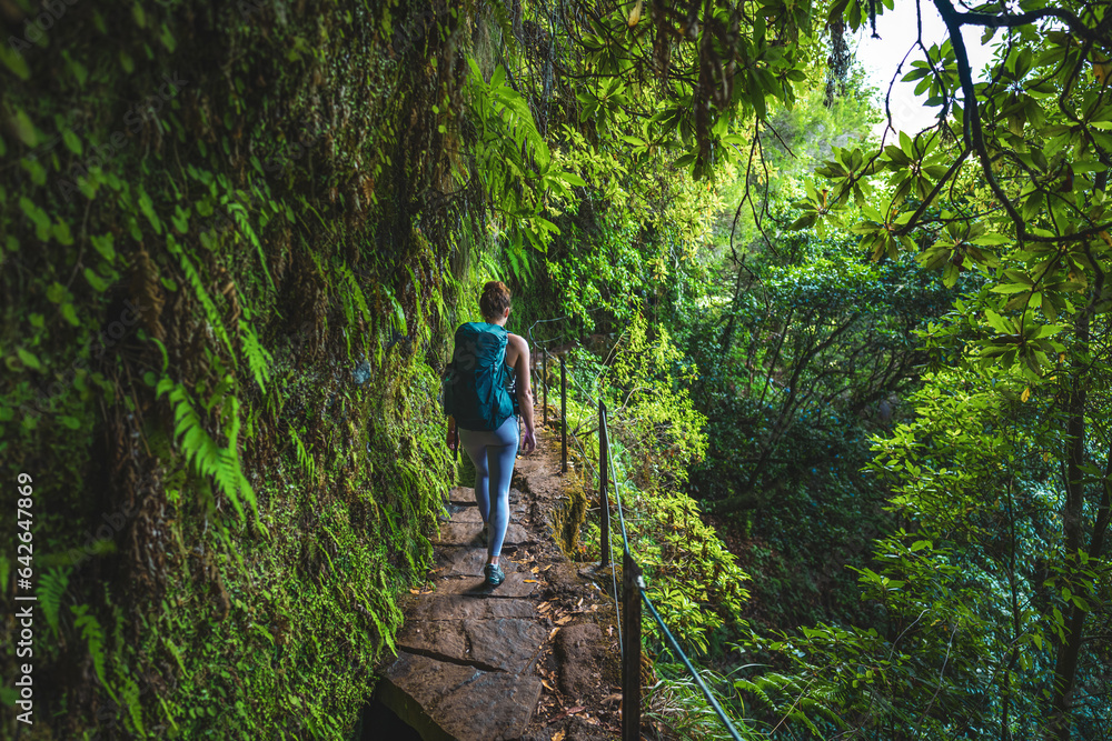Female backpacker walking along rainforest water channel path on steep cliff covered with plants. Levada of Caldeirão Verde, Madeira Island, Portugal, Europe. Female backpacker walking along rainforest water channel path on steep cliff covered with plants. Levada of Caldeirão Verde, Madeira Island, Portugal, Europe.