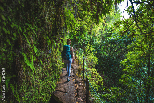 Female backpacker walking along rainforest water channel path on steep cliff covered with plants. Levada of Caldeirão Verde, Madeira Island, Portugal, Europe.