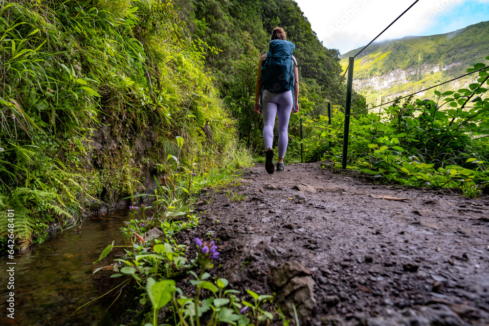 Low angle shot of backpacker walking along a picturesque hike trail under a cliff along a water channel through Madeira rainforest. Levada of Caldeirão Verde, Madeira Island, Portugal, Europe.