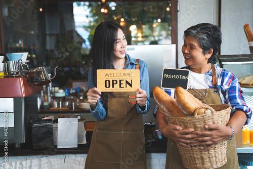 Portrait of a smiling senior Asian barista, hands hold bread basket with Promotion sign and smiling to her younger friend which hold Welcome Open sign.  Small business owner, family manage.
