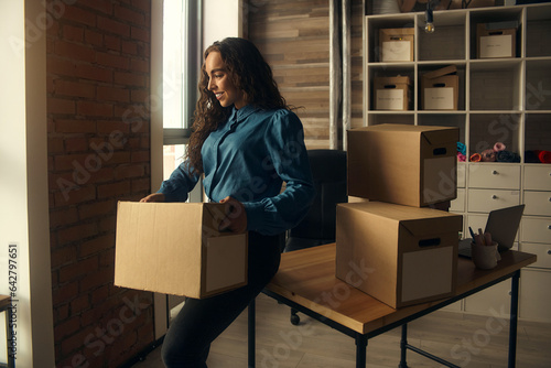 A young woman sits on a table in front of boxes, holding them in her hands, collecting parcels at the post office. entrepreneur online store