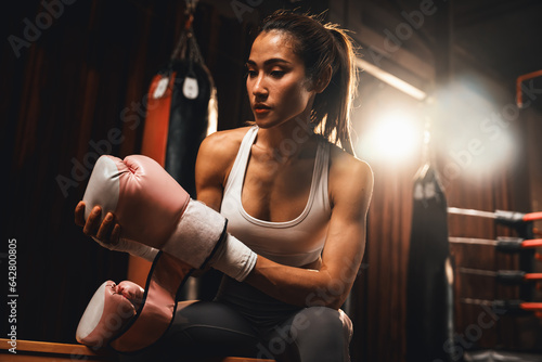 Determined Asian female Muay Thai boxer with muscularity physical readiness body wraps her hand and dons or wearing boxing glove, preparing for intense boxing training in the ring at gym. Impetus © Summit Art Creations