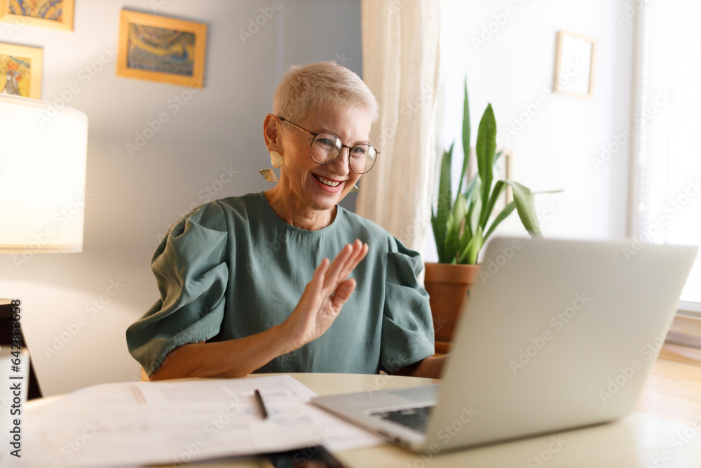 Senior trendy elegant female teacher in glasses with stylish haircut waving hand at camera of laptop saluting her students in beginning of online lesson, sitting at table of her home cabinet Senior trendy elegant female teacher in glasses with stylish haircut waving hand at camera of laptop saluting her students in beginning of online lesson, sitting at table of her home cabinet