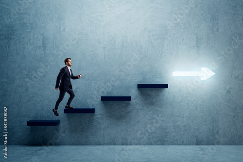 Side view of young business man climbing illuminated arrow stairs to success on concrete wall background in interior. Financial growth, career development and forward concept.