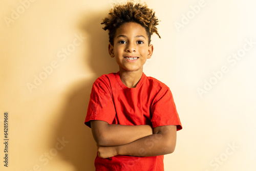 An African boy between 5 and 6 years old with afro hair poses on a brown studio background with a red T-shirt. The little one is with his arms crossed looking at the camera happily.